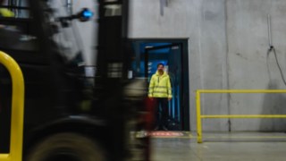 En man i säkerhetskläder stiger ut ur en dörr i en fabrikshall. En truck kör förbi. En man i säkerhetskläder stiger ut ur en dörr i en fabrikshall. En truck kör förbi.
