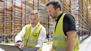 Parallella laddningsprocesser kan leda till oönskade effekttoppar. Linde connect:charger löser problemet. Två män i gula varselvästar står i ett höglager och tittar på en laptop.