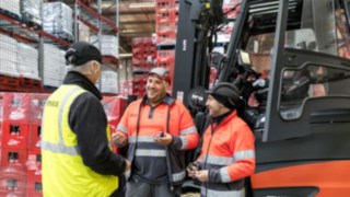 Komforten och säkerheten i truckarna från Linde höjer förarens arbetsnöjdhet och motivation. Tre personer pratar framför en truck.